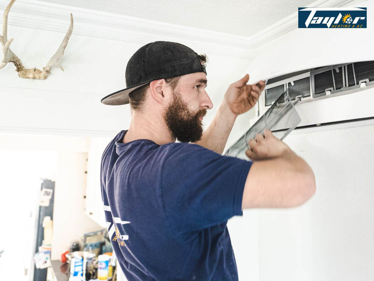 Bearded man in blue shirt installing or replacing an air conditioning filter in a residential HVAC unit mounted on wall.