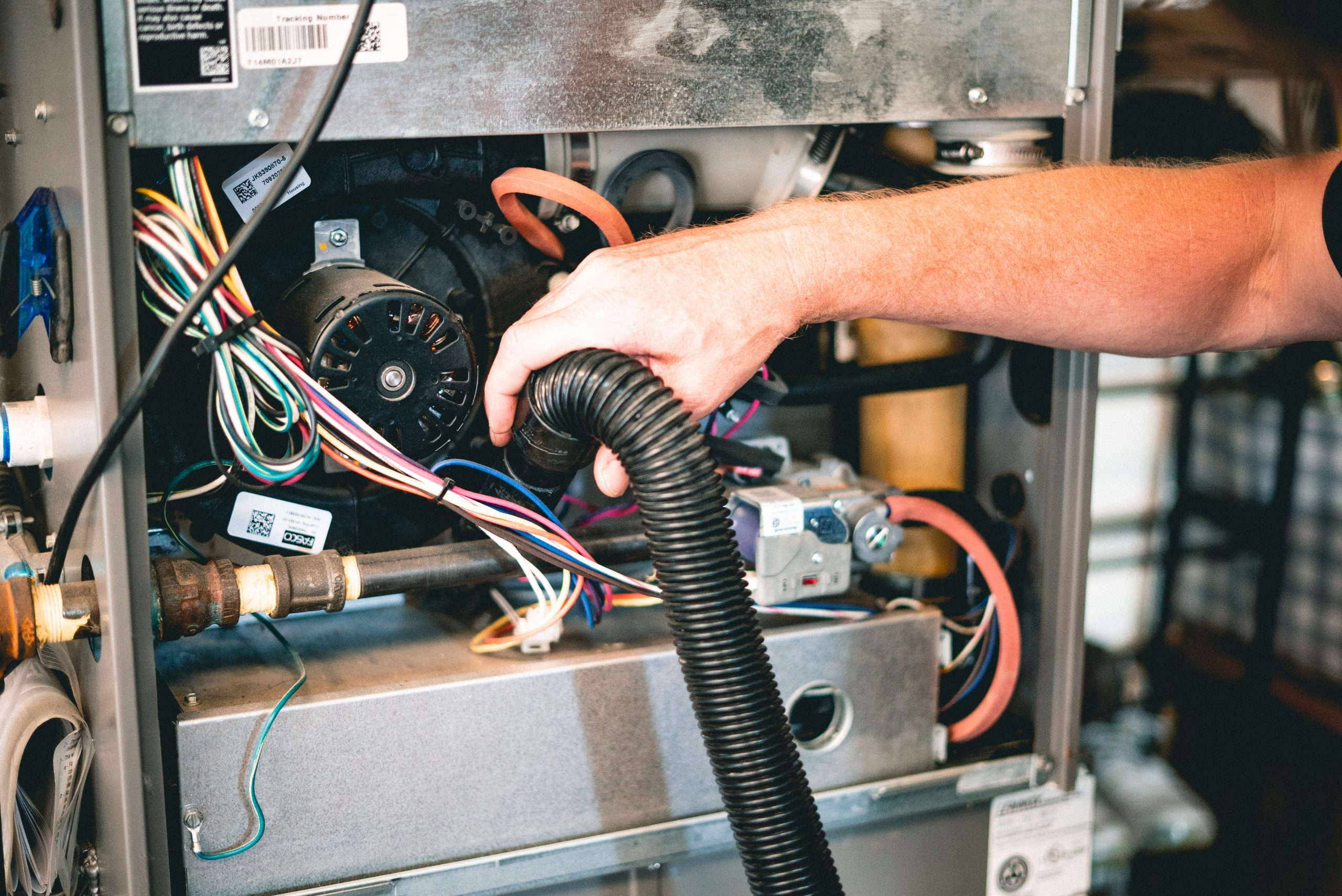 Technician's hand holding a black ribbed duct hose inside an HVAC furnace unit with visible wiring, motor, and metal components.