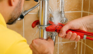 Plumber in yellow shirt tightening red valve on burst water pipe with wrench in bathroom.
