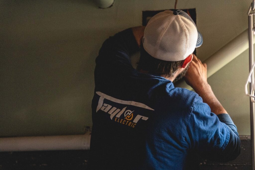 Technician wearing white cap and Taylor Electric branded shirt working on ceiling installation indoors.