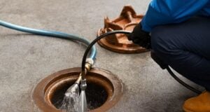 Worker using a high-pressure water hose to clean an open sewer drain pipe on concrete ground.