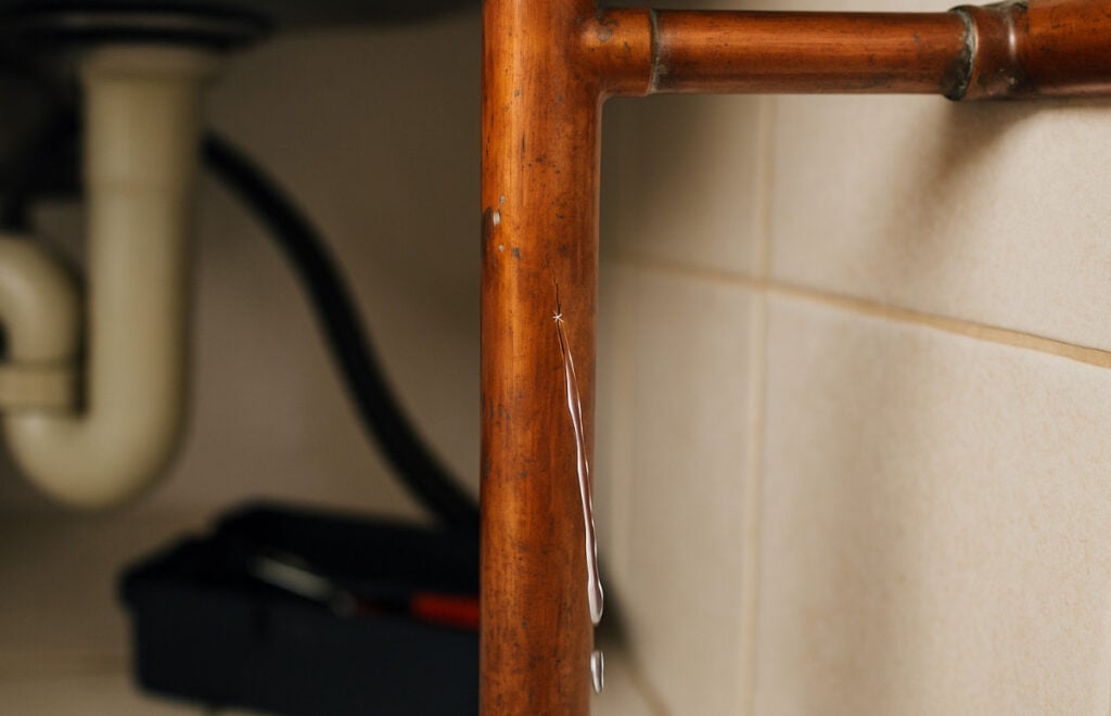 Water dripping from a crack in a copper pipe with visible damage and patina, against a tiled wall background.