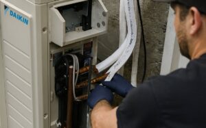 HVAC technician wearing blue gloves inspects internal components of a DAIKIN heat pump system with exposed copper piping and white refrigerant lines.