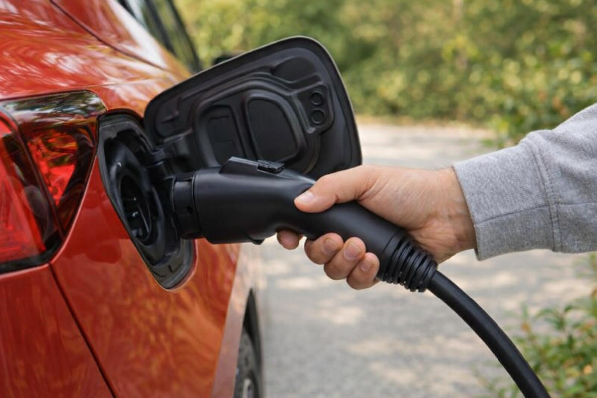 Person holding black electric vehicle charging cable connected to red car's charging port outdoors.