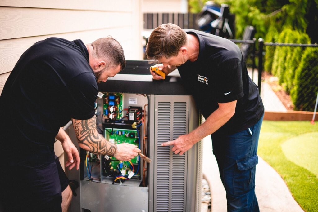 Two technicians in black shirts inspect internal components of an outdoor HVAC air conditioning unit in a residential backyard.