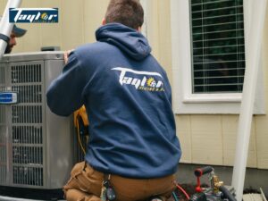 HVAC technician wearing Taylor Heating & AC branded navy hoodie performing maintenance on an air conditioning unit outdoors.