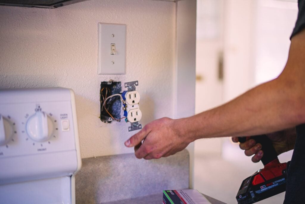 Electrician's hands installing a white duplex outlet into wall box with switch plate and wiring visible above.