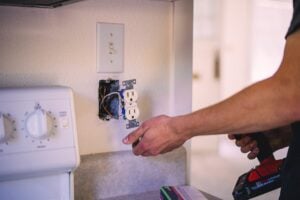 Electrician's hands installing a white duplex outlet into wall box with switch plate and wiring visible above.
