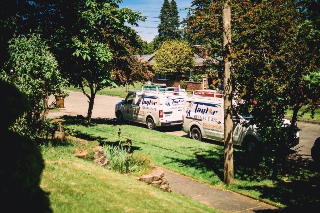 Two white Taylor Heating & A/C service vans parked on a tree-lined residential street with brick homes visible in background.