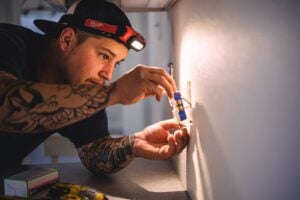 Tattooed electrician wearing Milwaukee headlamp installs electrical outlet in drywall opening.