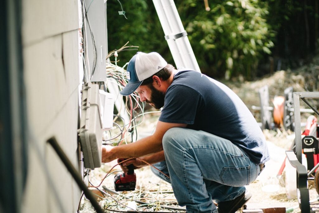 Bearded electrician wearing blue cap crouched down working on outdoor electrical wiring and connections on white panel.
