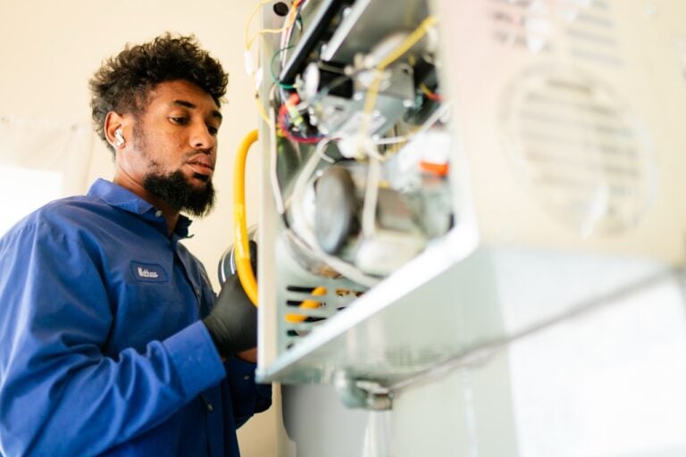 Male technician in blue work shirt inspecting an open electrical panel with colorful wiring and components.