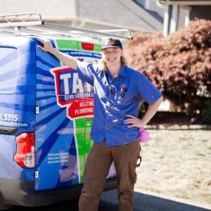 Smiling service technician in blue uniform standing beside a branded heating and plumbing company van in a residential driveway.