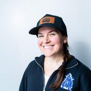 Woman wearing a navy baseball cap with orange TAYLOR patch, dark zip-up hoodie with blue emblem, smiling at camera against white background.