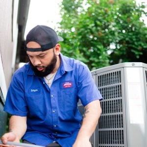 Technician in blue Johnson service uniform inspecting outdoor AC condenser unit in residential setting.